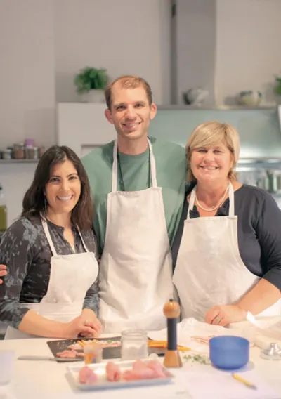 Three smiling cooks in white aprons at a kitchen counter with sliced ingredients before them