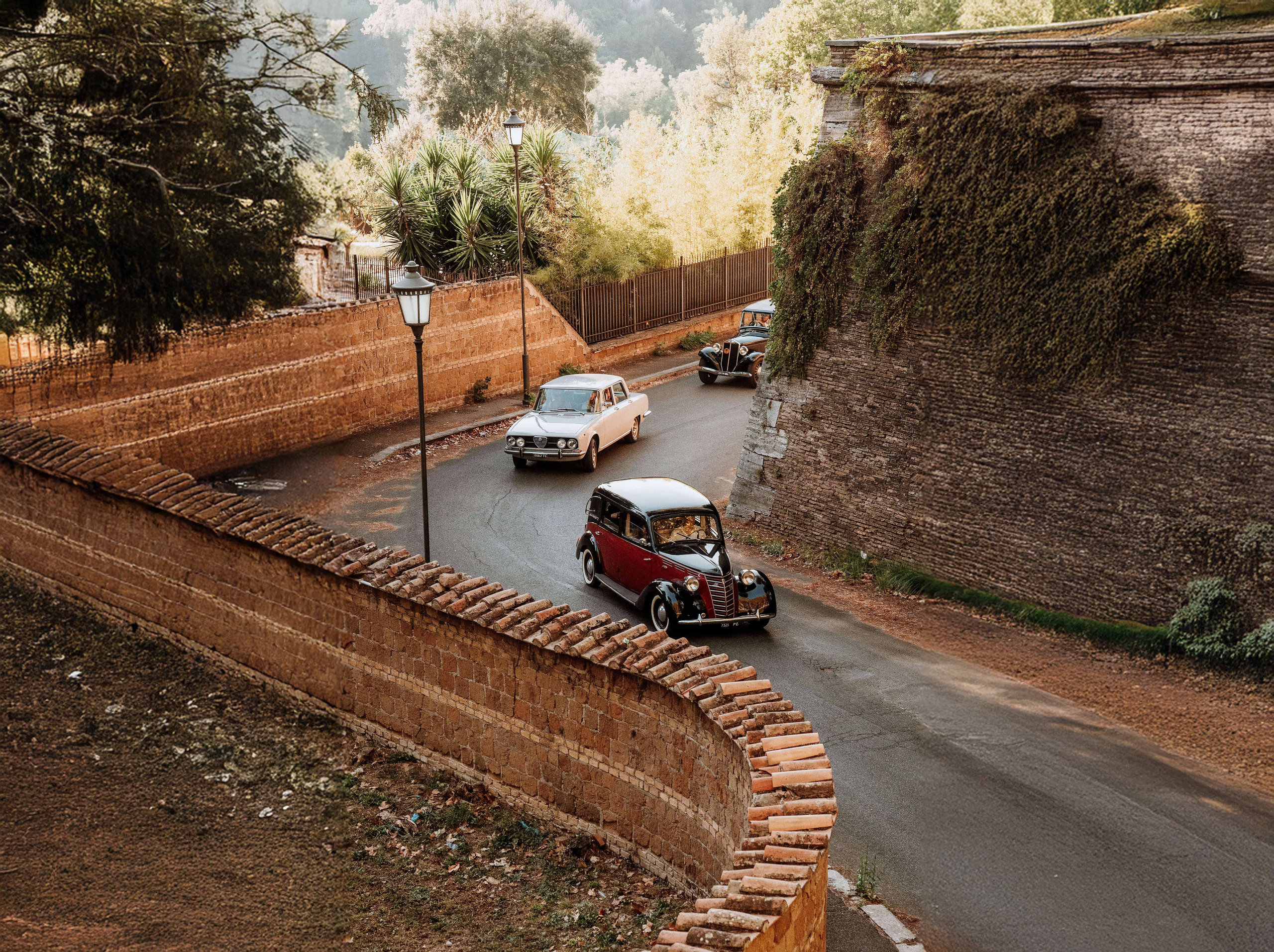 Vintage cars driving along a curving road beside old brick walls and tall trees
