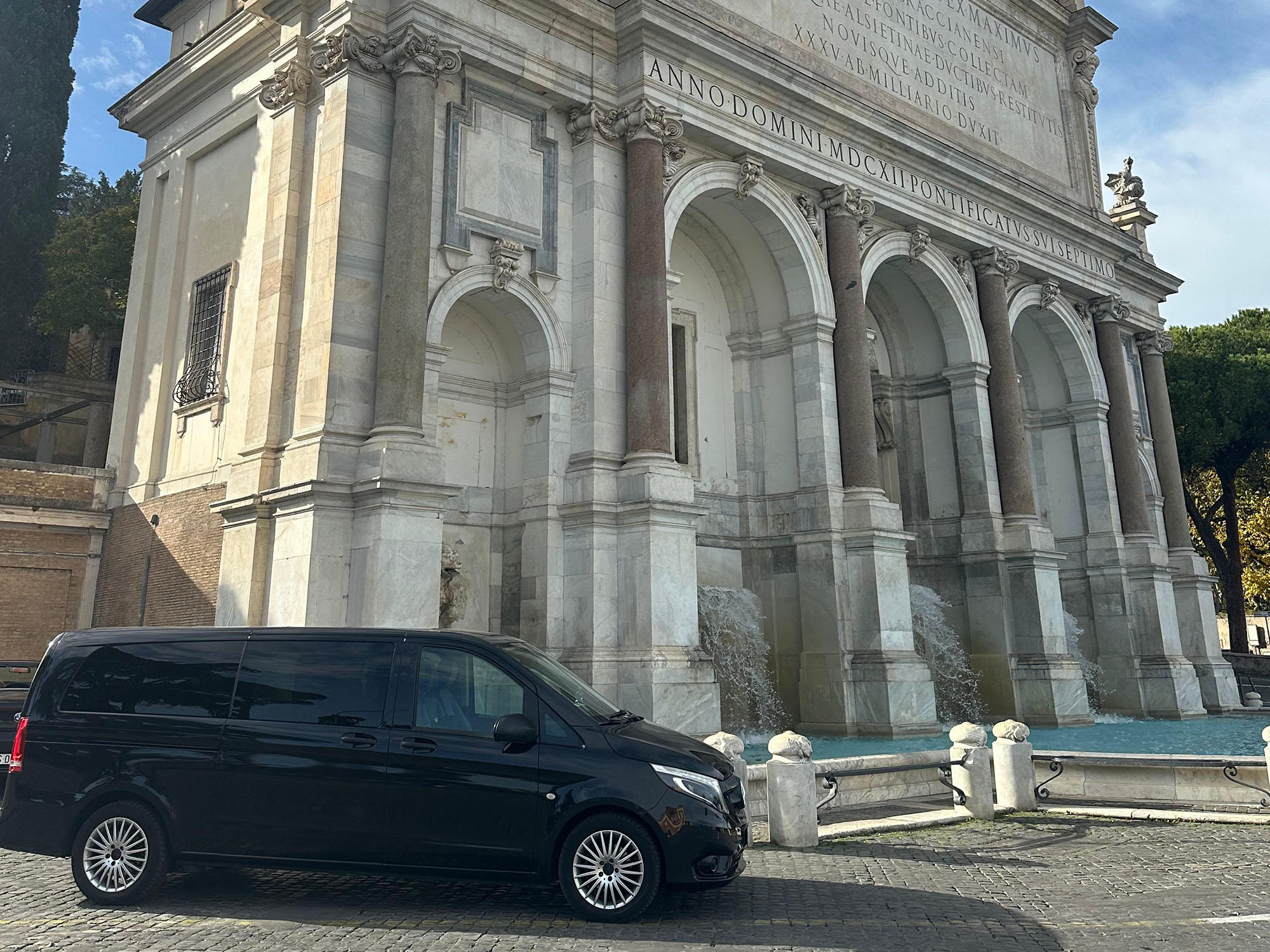 Black executive minivan parked on a cobbled square in front of a monumental Roman fountain
