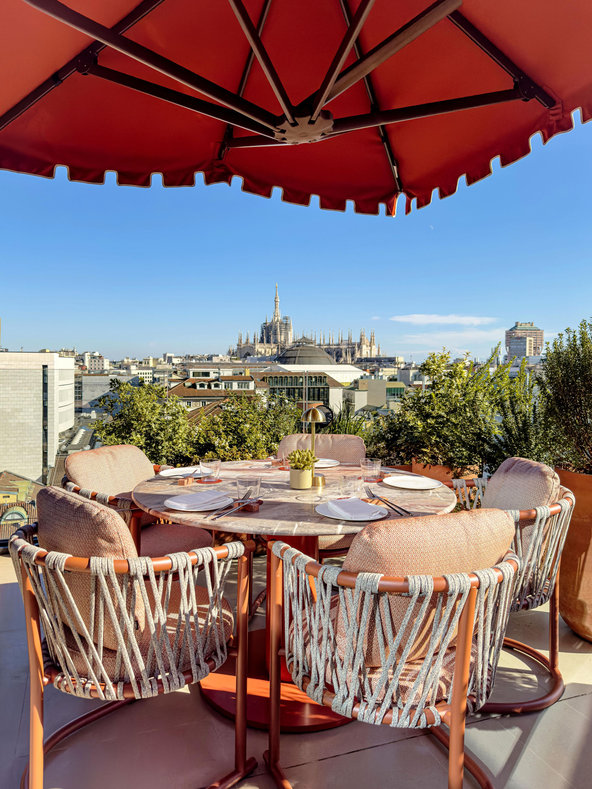 A rooftop restaurant table under a red parasol, the spires of the Duomo in the distance