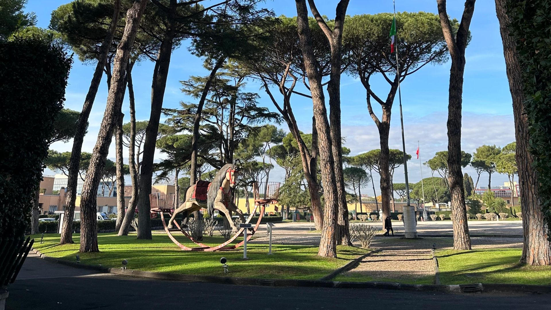 Large wooden rocking horse on a lawn among tall umbrella pines with an Italian flag in the distance