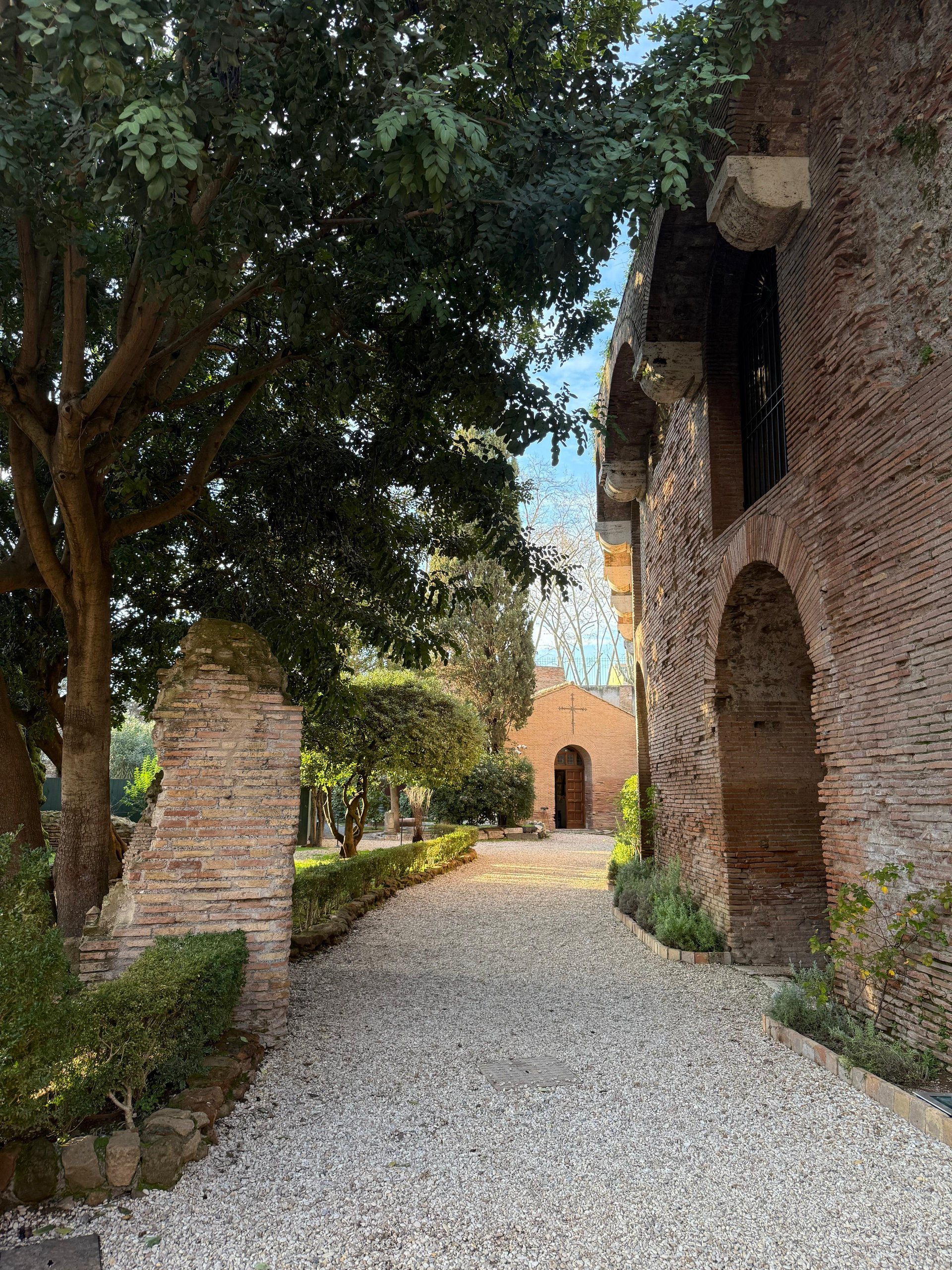 A gravel path between brick walls and trees in the park above the Domus Aurea