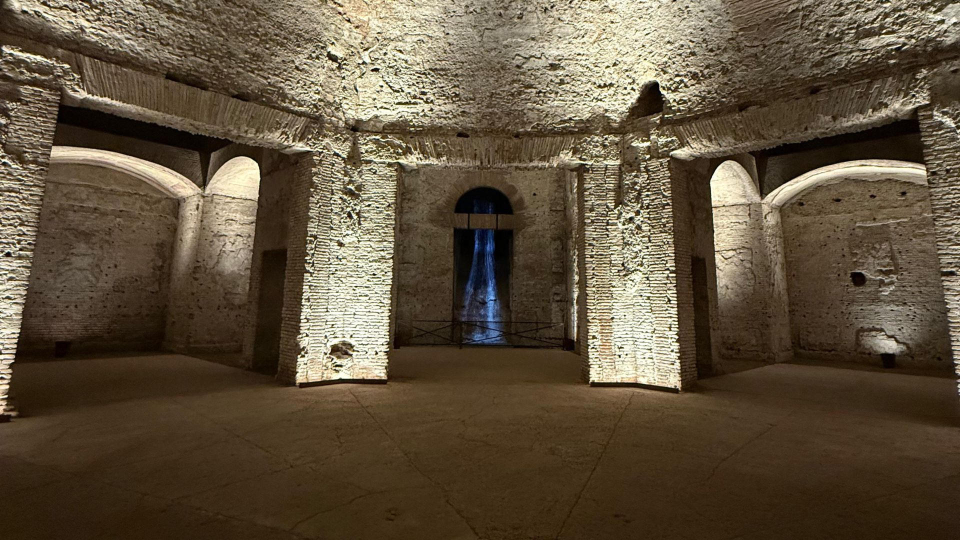 Vaulted octagonal hall of brick and stone with a beam of blue light at the far end of an underground Roman palace