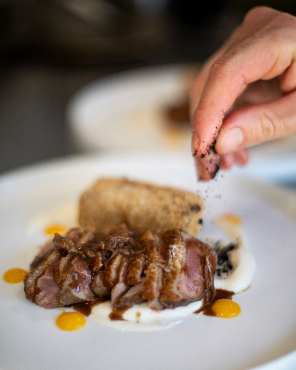 A chef finishing a plated meat course with a sprinkle of seasoning