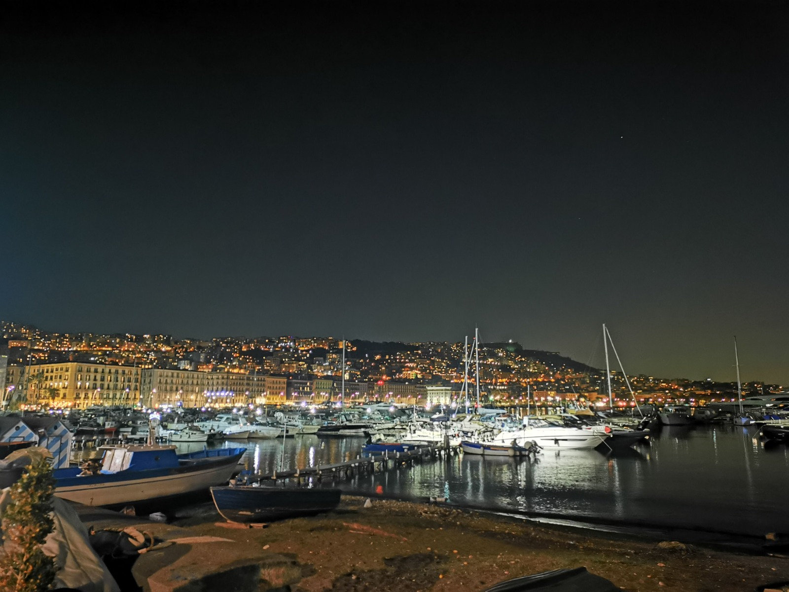 Bay of Naples marina at night with moored boats and the lit hillside city reflected in the water