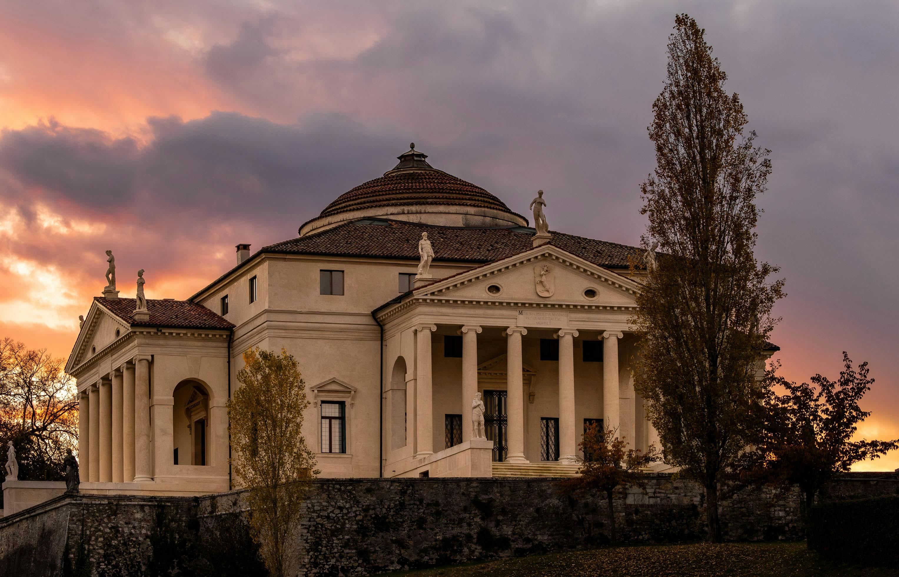 Neoclassical villa with a columned portico and shallow dome glowing against a pink and purple sunset sky