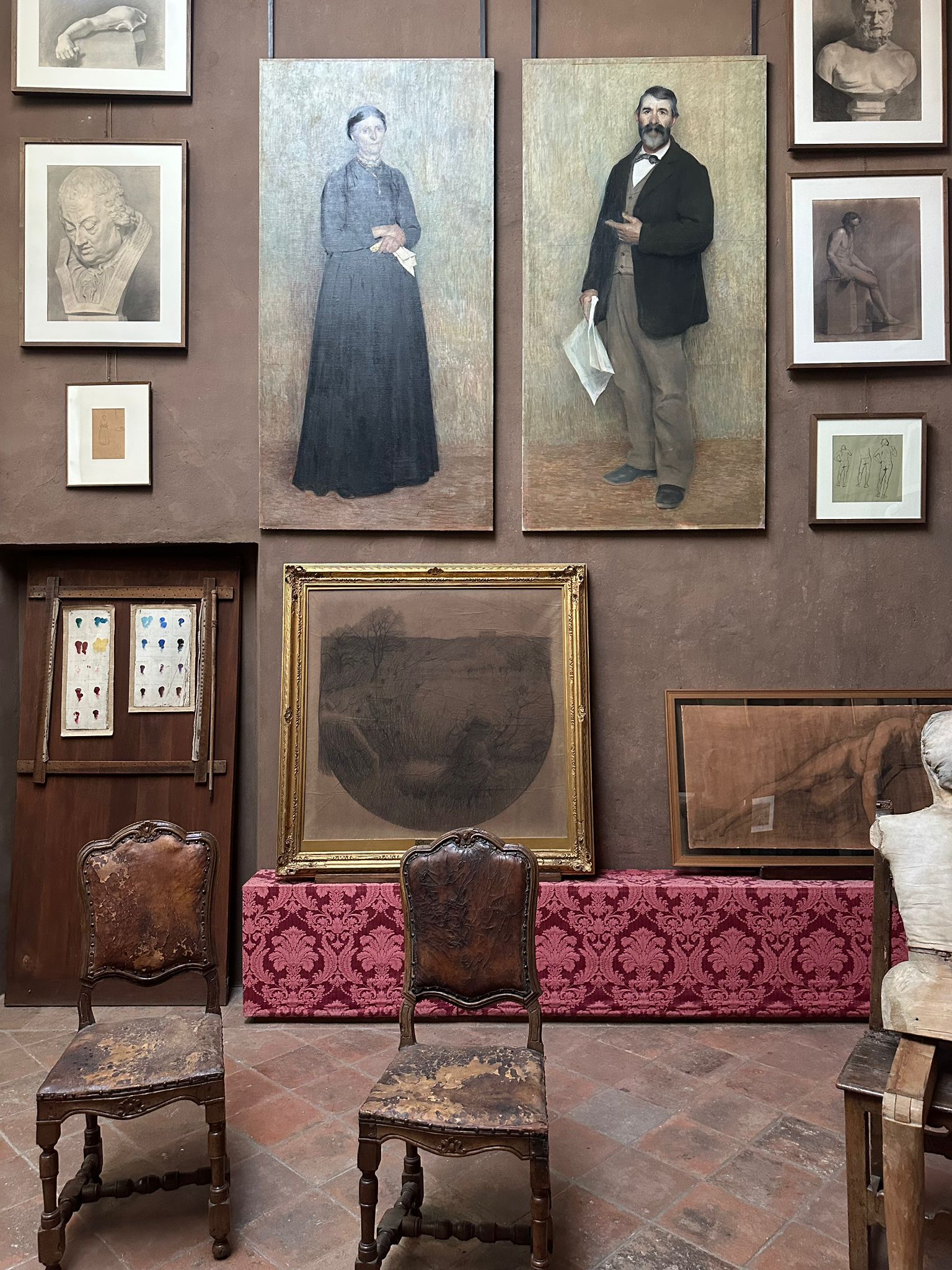 Artist's studio wall hung with full-length portraits, sketches and plaster busts above two antique leather chairs