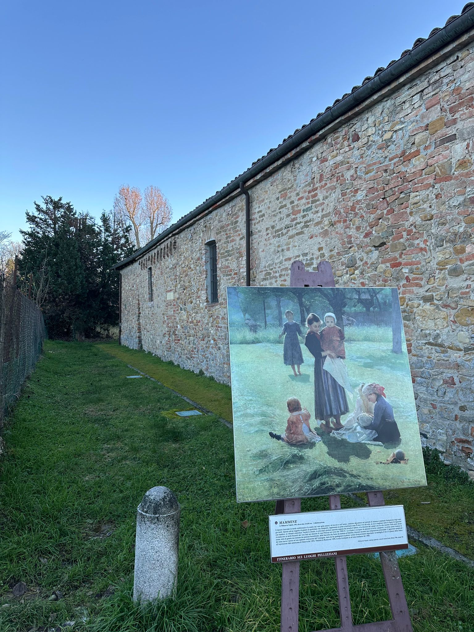 Painting reproduction on an easel in the grass beside a long brick farm building under a clear sky