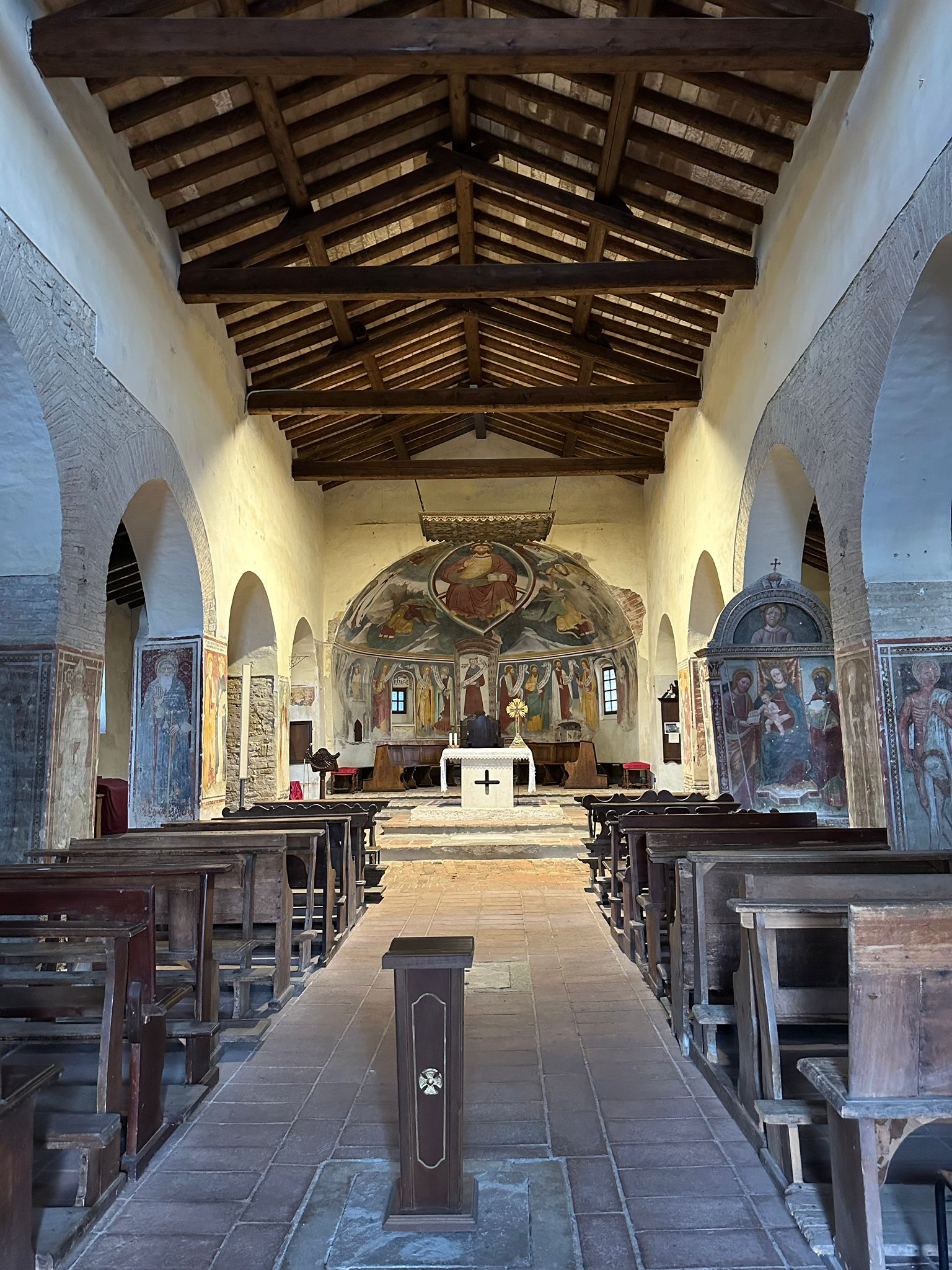 Nave of a small country church with wooden beams, pews and a frescoed apse behind the altar