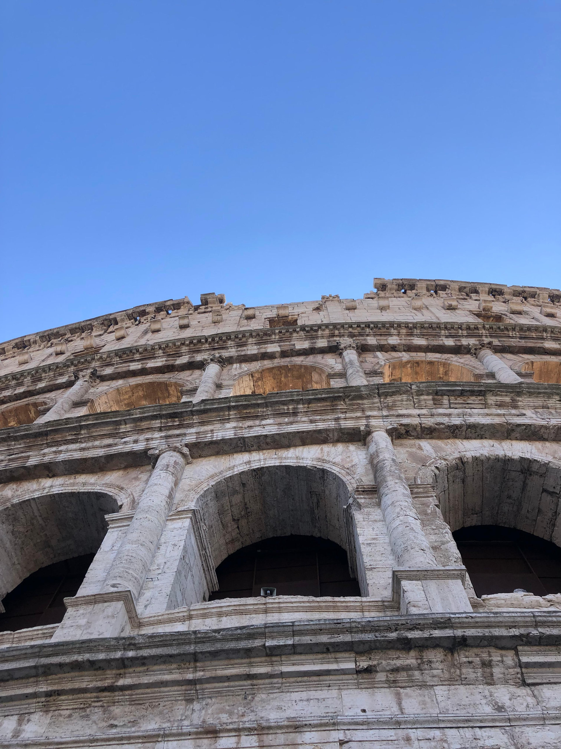 Looking up at the weathered travertine arches and tiers of the Colosseum against a clear blue sky