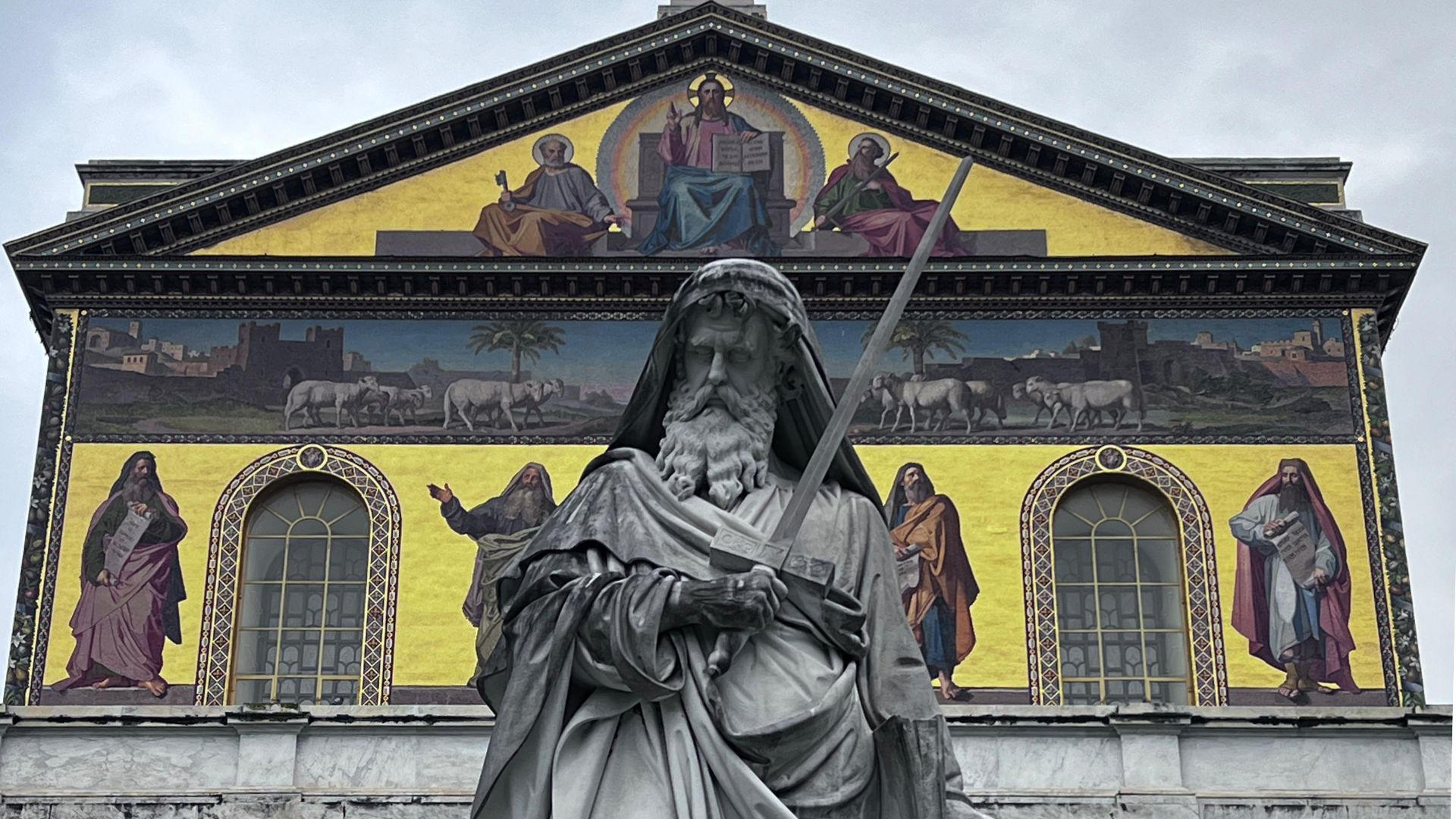 Statue of Saint Paul with a sword in front of the gold mosaic facade of a Roman basilica