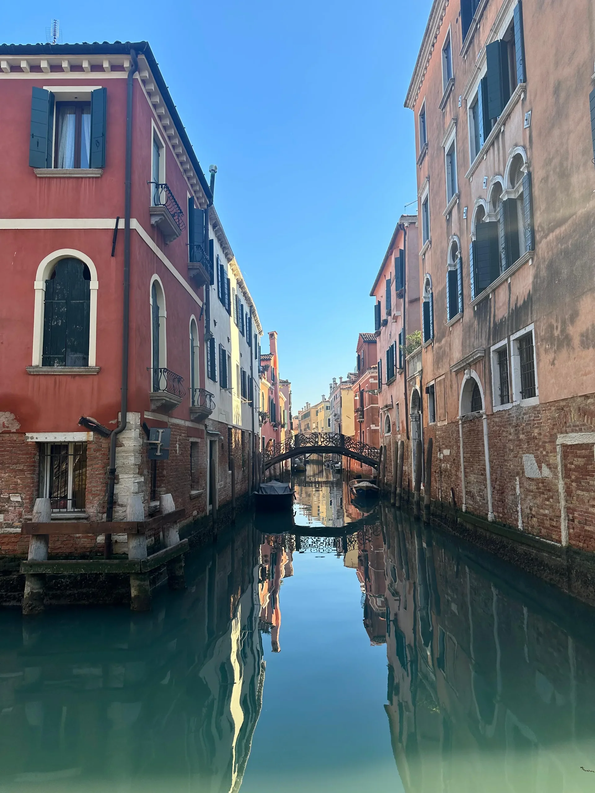 Quiet Venetian canal lined with red and ochre houses, with a small bridge and reflections in the green water