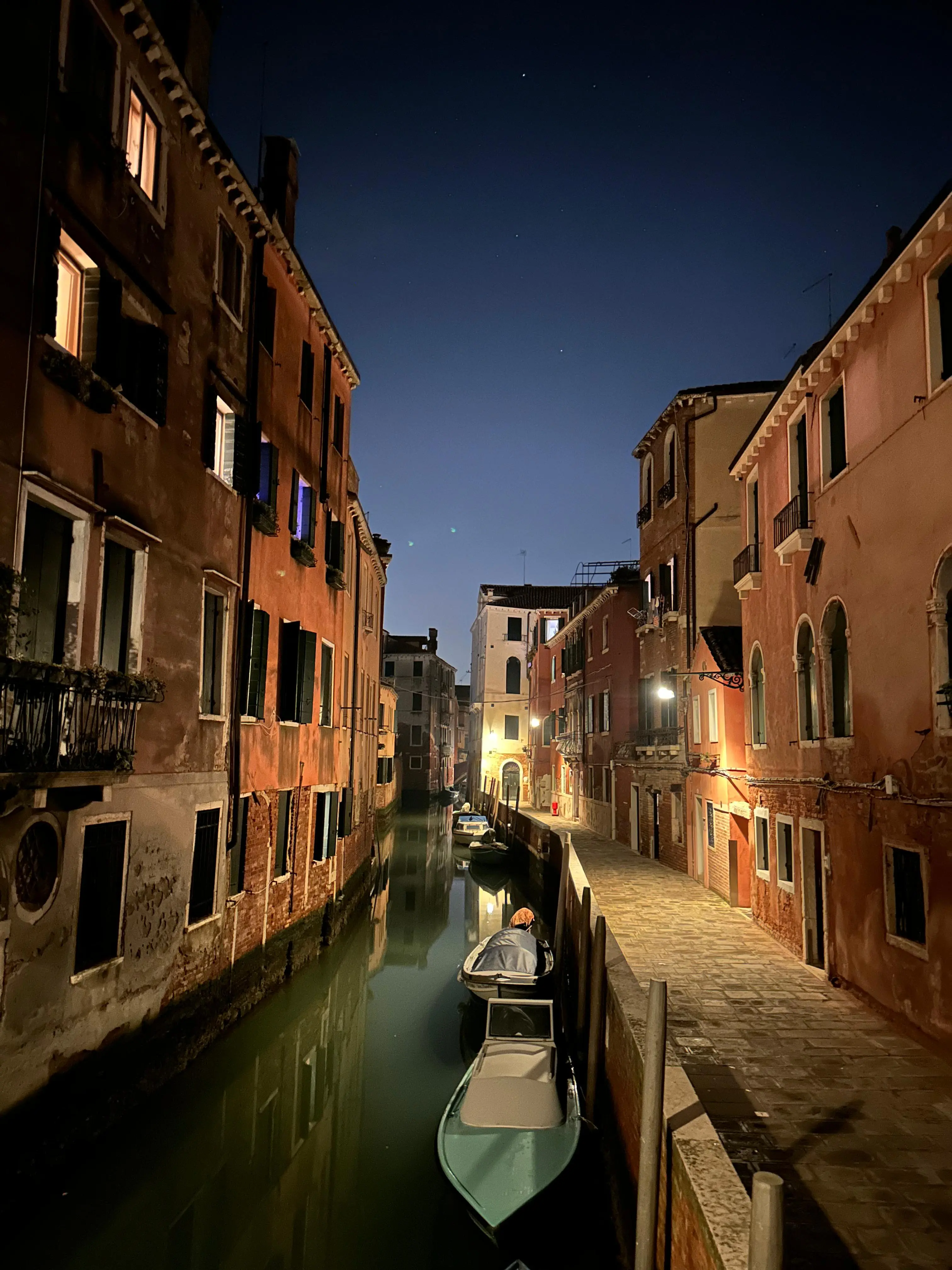 Quiet Venetian canal at night, moored boats and lit windows along a stone walkway
