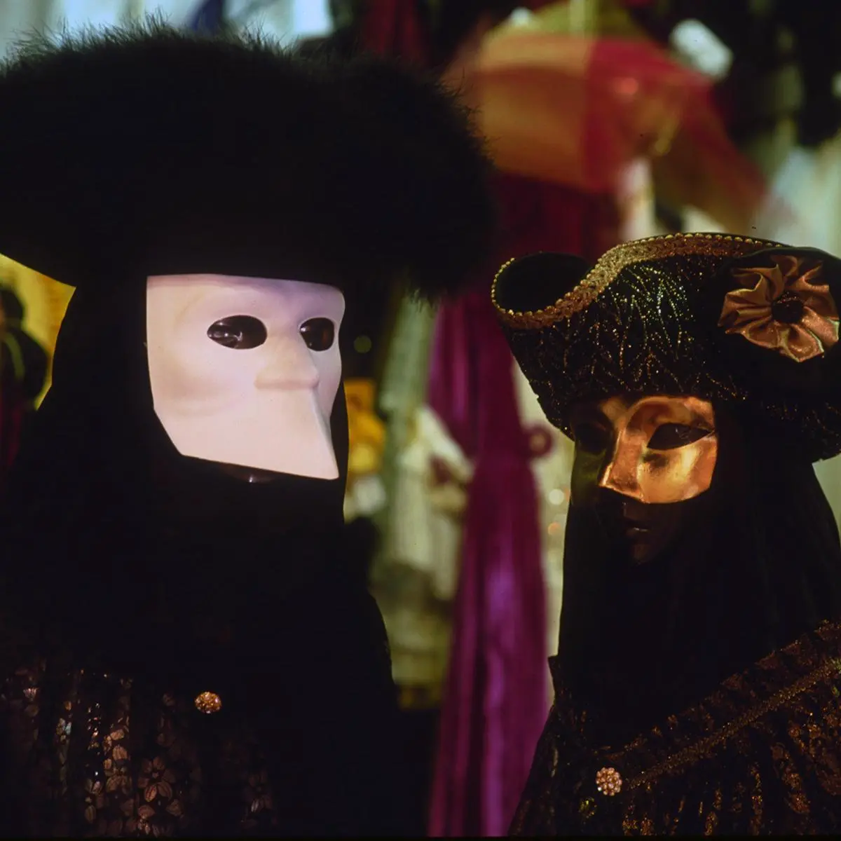 Two Carnival figures in black capes and tricorn hats, one in a white bauta mask, one in gold