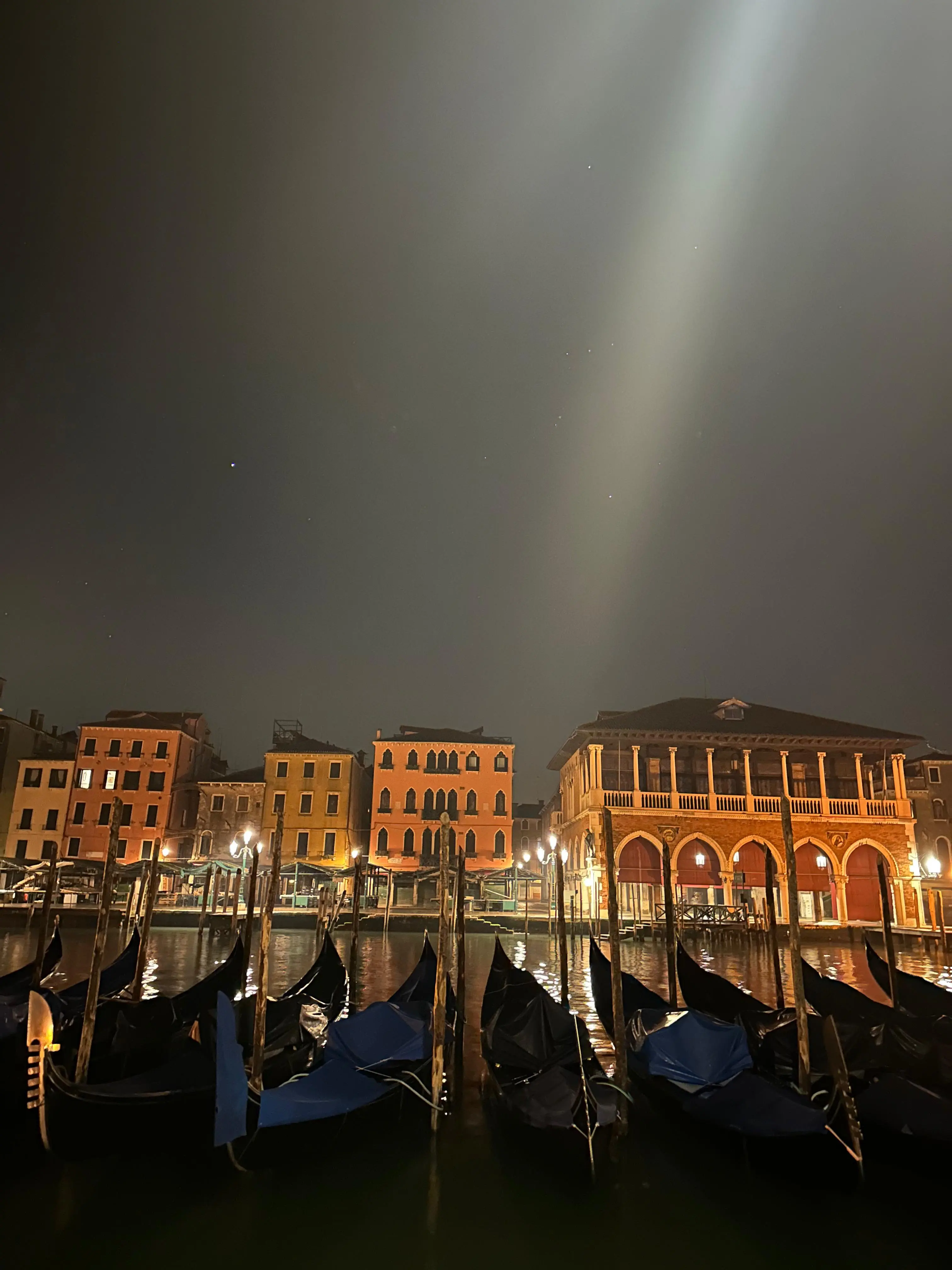 Covered gondolas moored on the Grand Canal at night, with lit palazzi and a beam of light in the sky