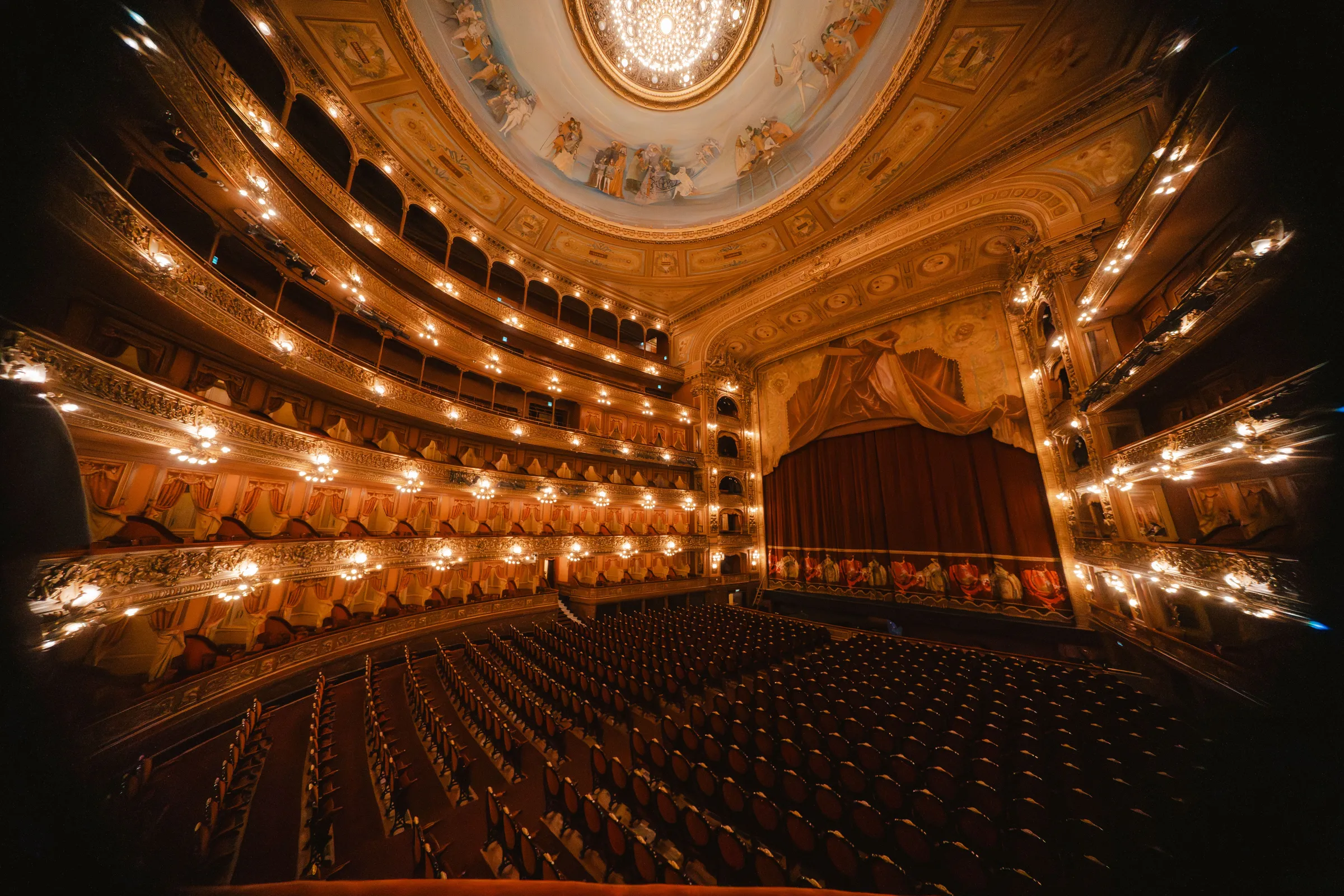 Ornate opera house interior with tiered gilded balconies, a frescoed dome and rows of empty seats