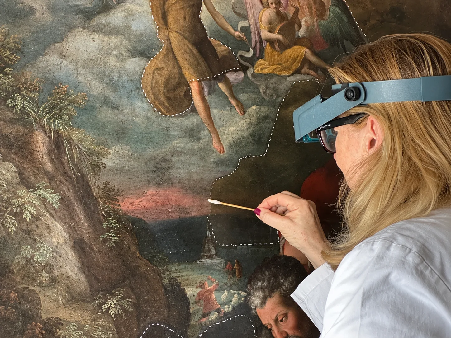 A restorer wearing a magnifying visor cleans an old oil painting of angels and shepherds with a cotton swab