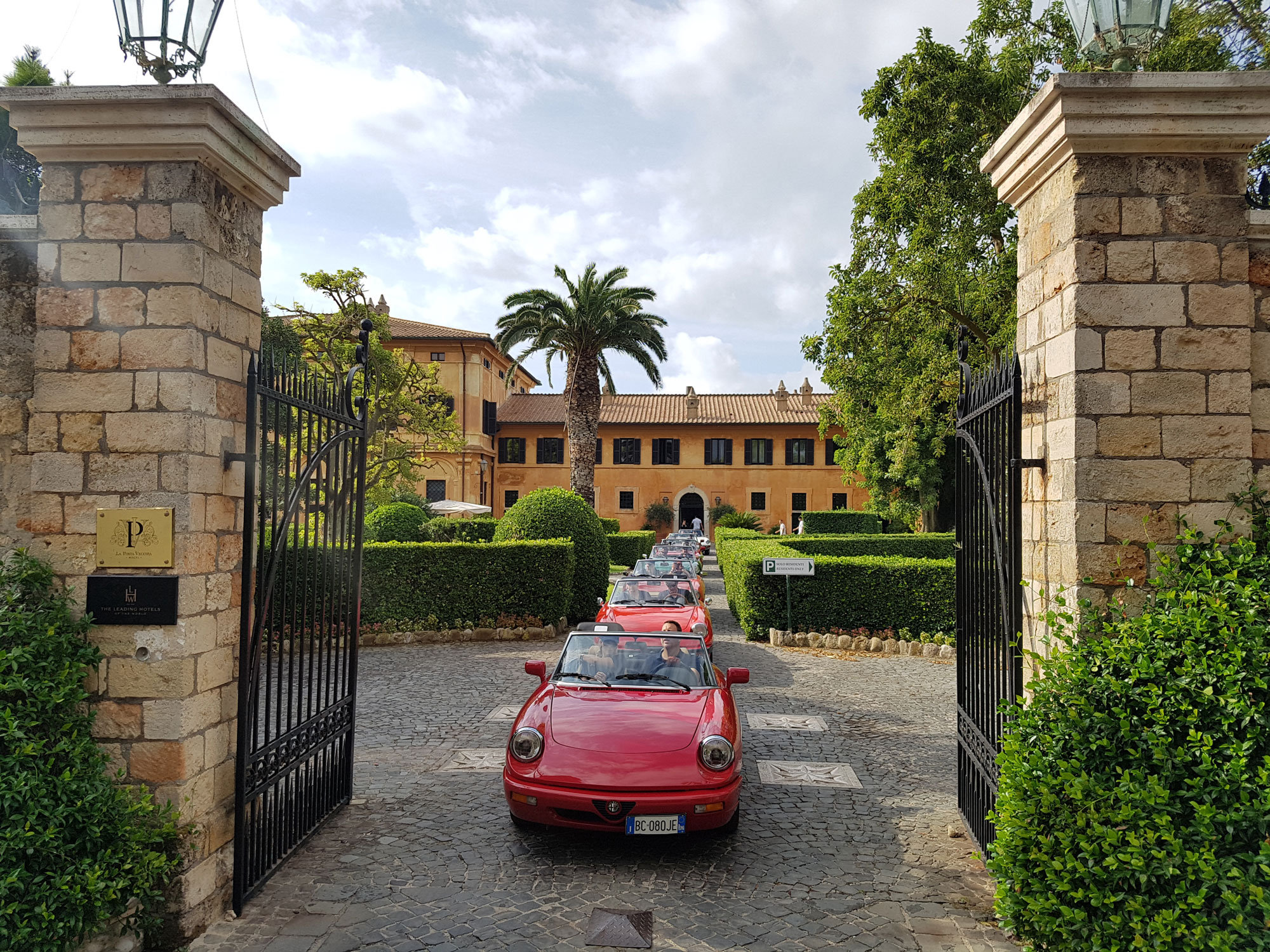 Convoy of red vintage sports cars driving through a stone gateway towards an ochre villa