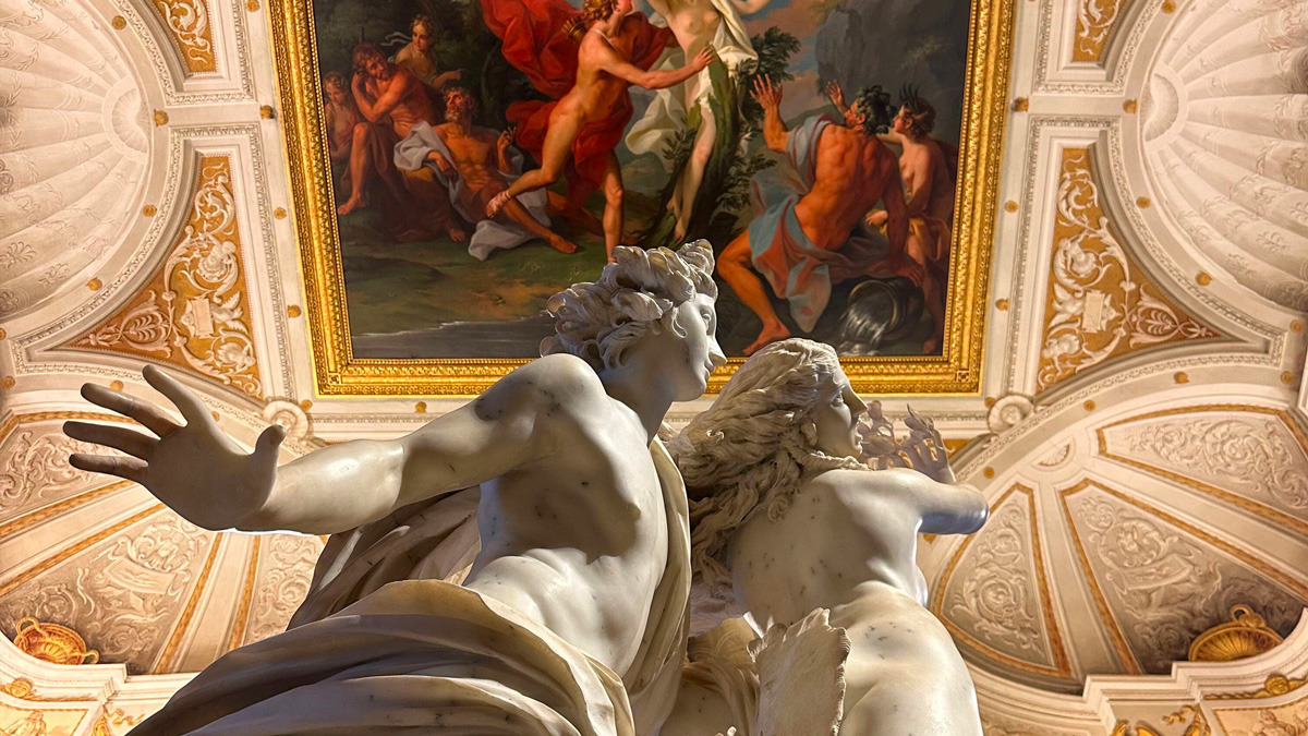 Marble sculpture of a chase between two figures beneath a gilded, frescoed ceiling in the Borghese Gallery