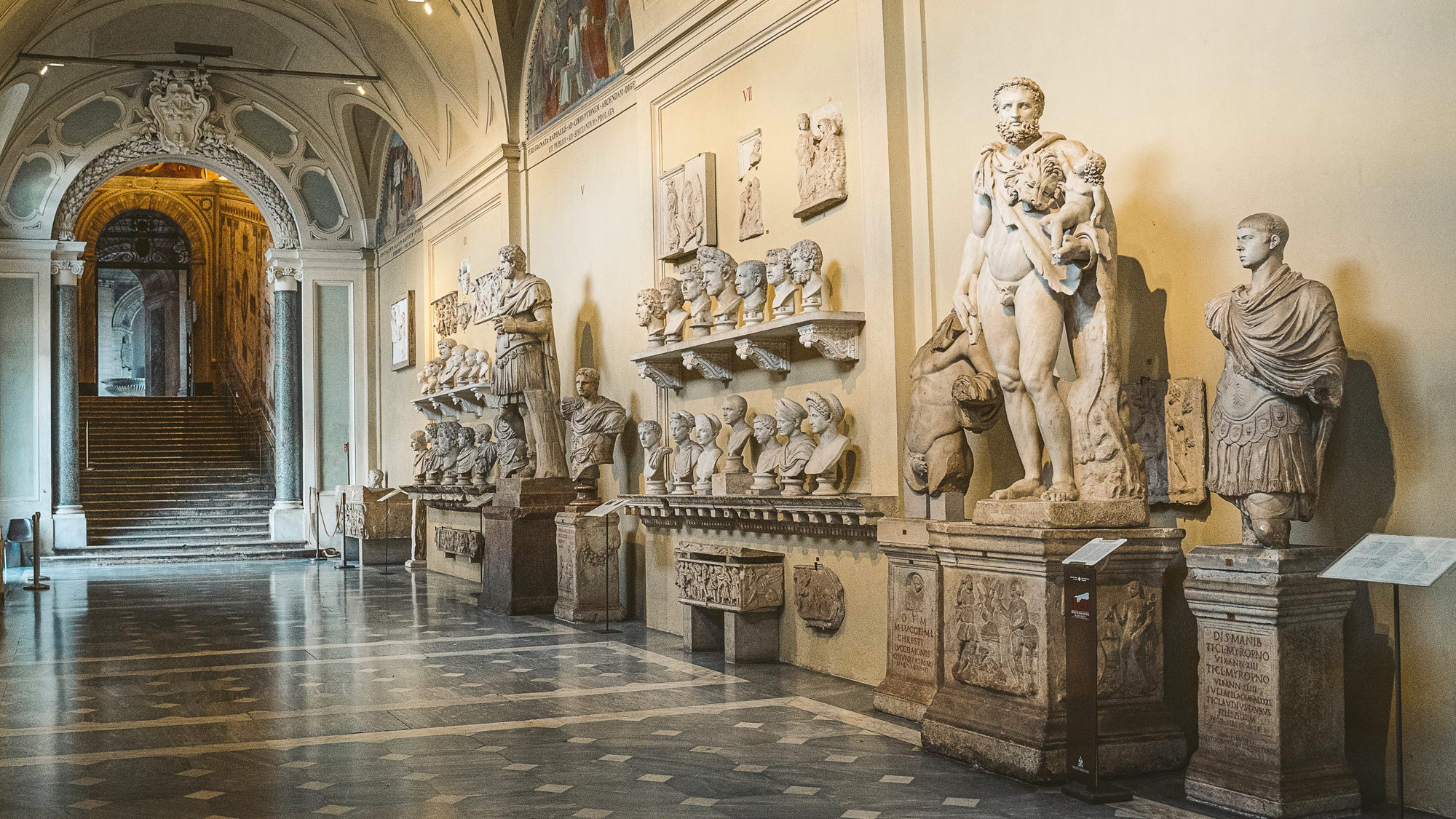 Vatican Museums gallery of ancient Roman statues and busts with a grand staircase beyond