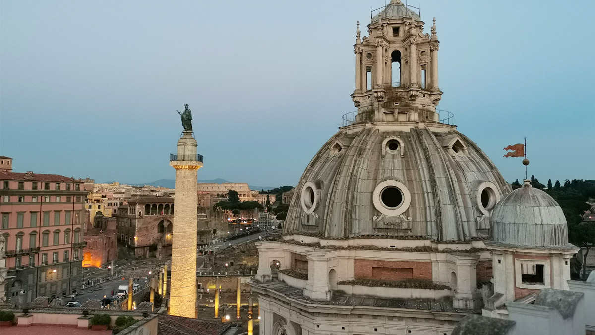 Trajan's Column and a Baroque church dome at dusk, with the city lights coming on