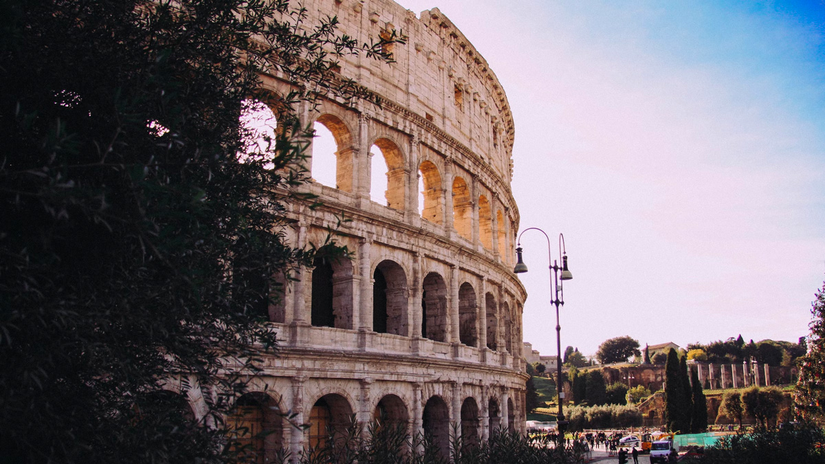 Curving outer wall of the Colosseum in warm evening light, framed by dark foliage