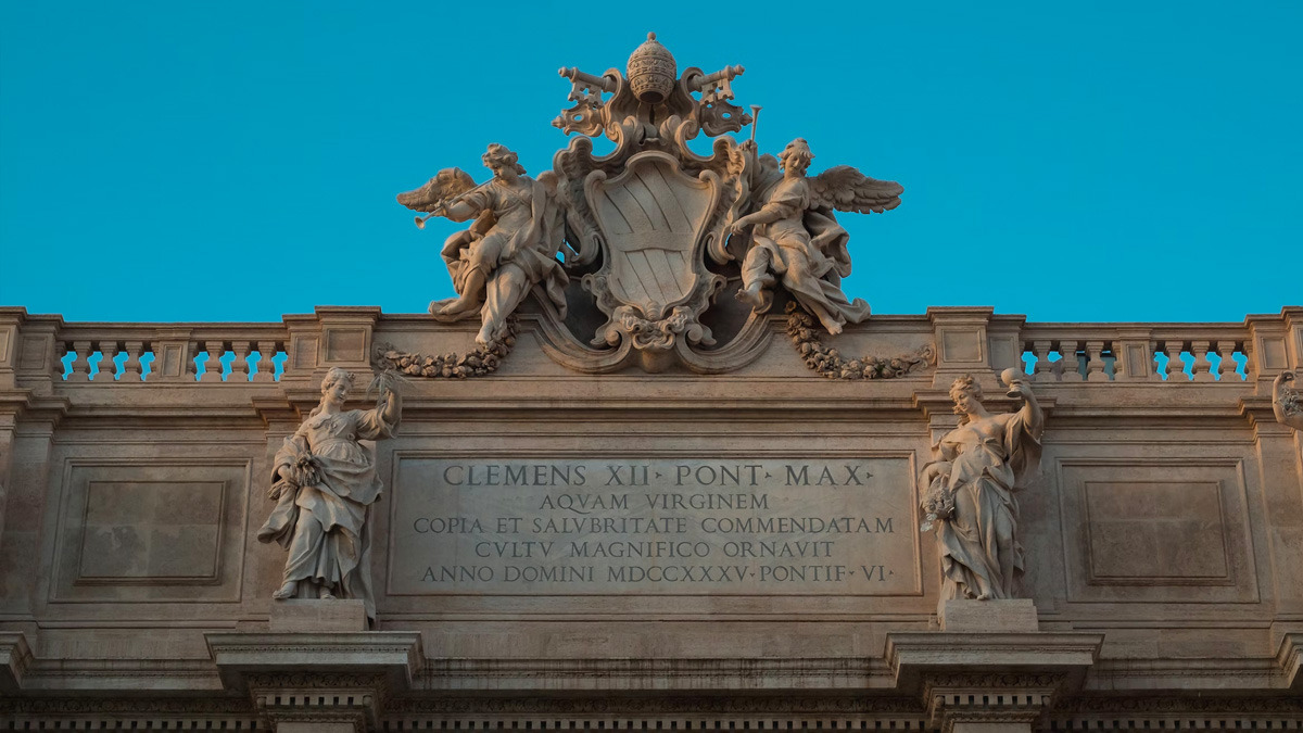 Top of the Trevi Fountain facade with angels, papal coat of arms and Latin inscription against blue sky