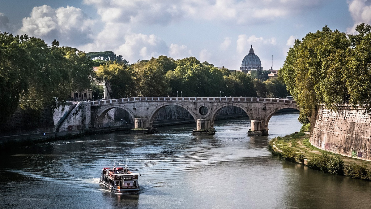 Tourist boat on the Tiber beneath a stone bridge with the dome of St Peter's in the distance