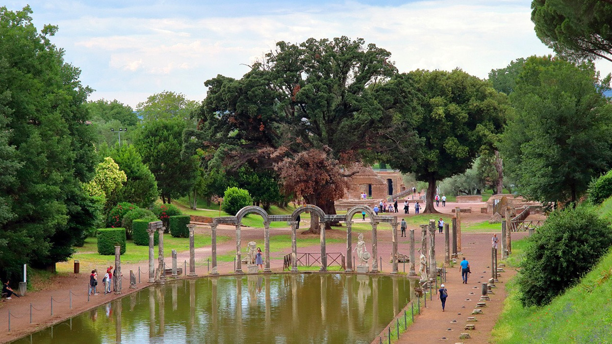 Visitors strolling beside the long pool of the Canopus among green trees and statues