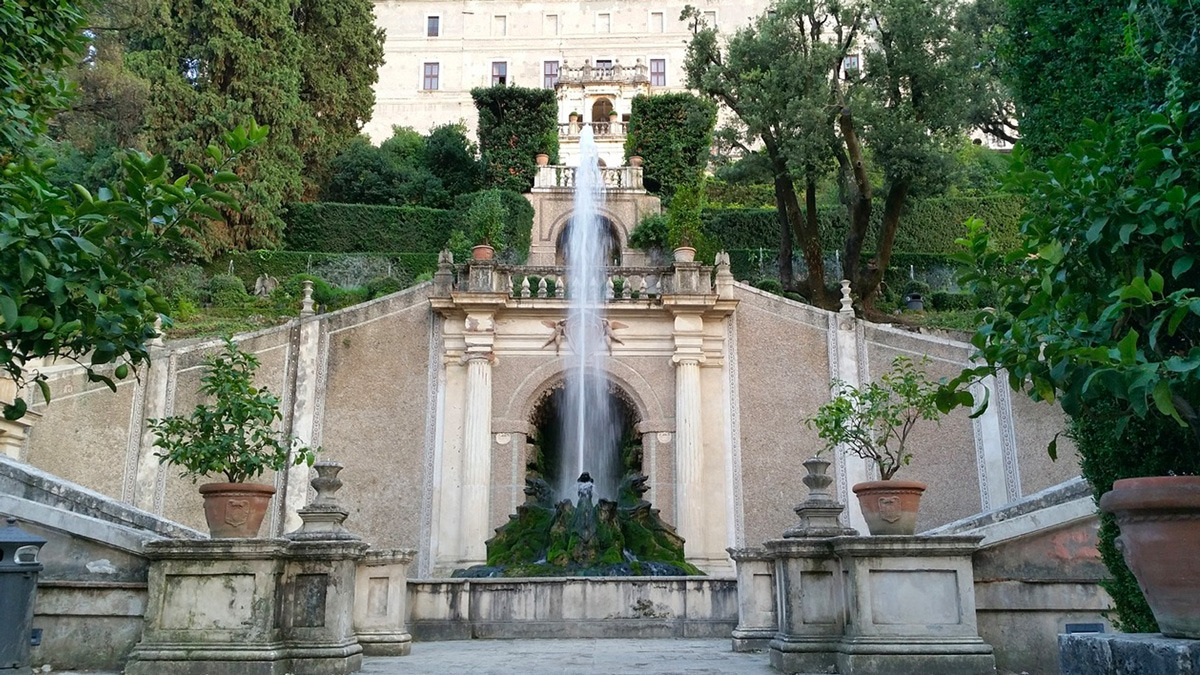 Small garden fountain beneath an arched stone niche, with potted citrus trees and a palace behind