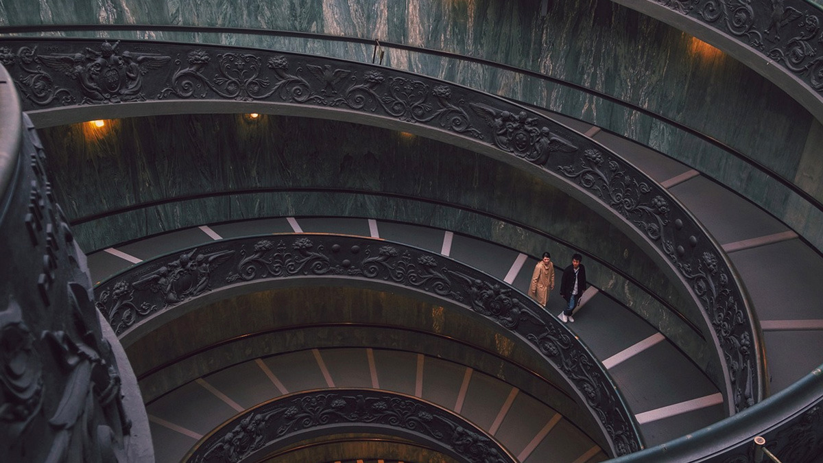 Two visitors walking down the ornate bronze double-helix spiral staircase of the Vatican Museums