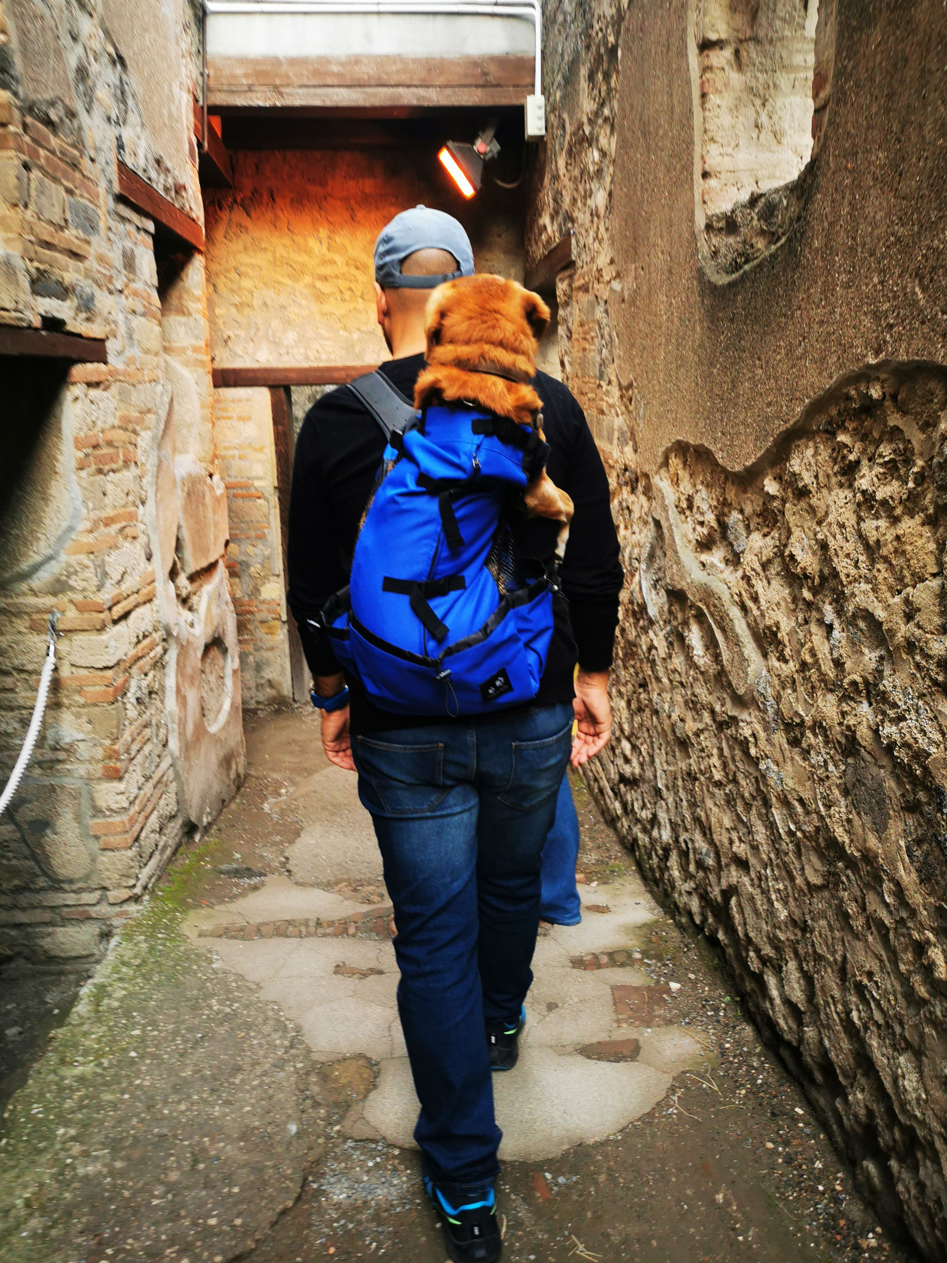 A visitor carrying a dog in a blue backpack along a narrow passage between ancient stone walls