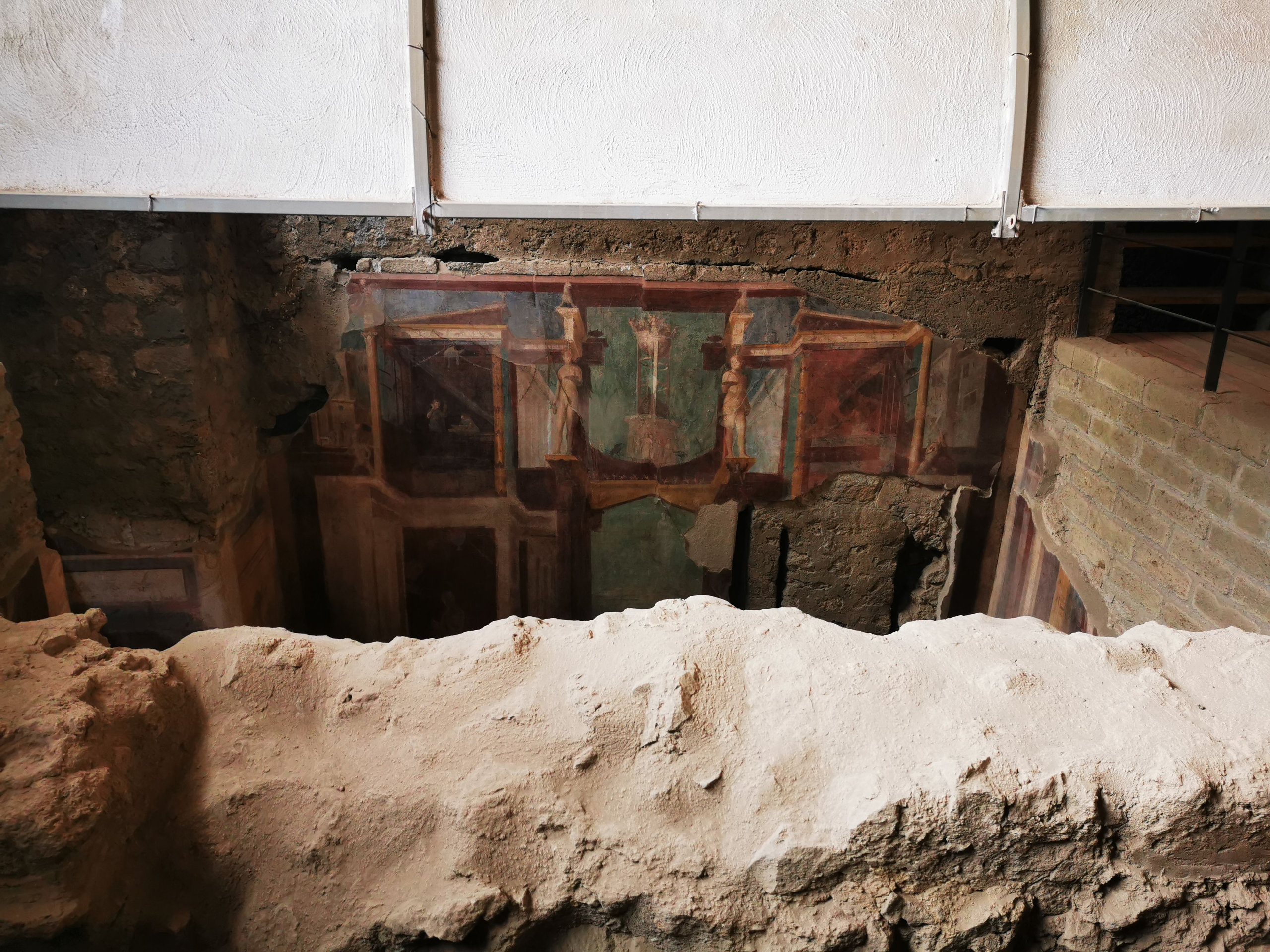 Painted wall decoration seen through a gap above a protective canopy in an excavated house