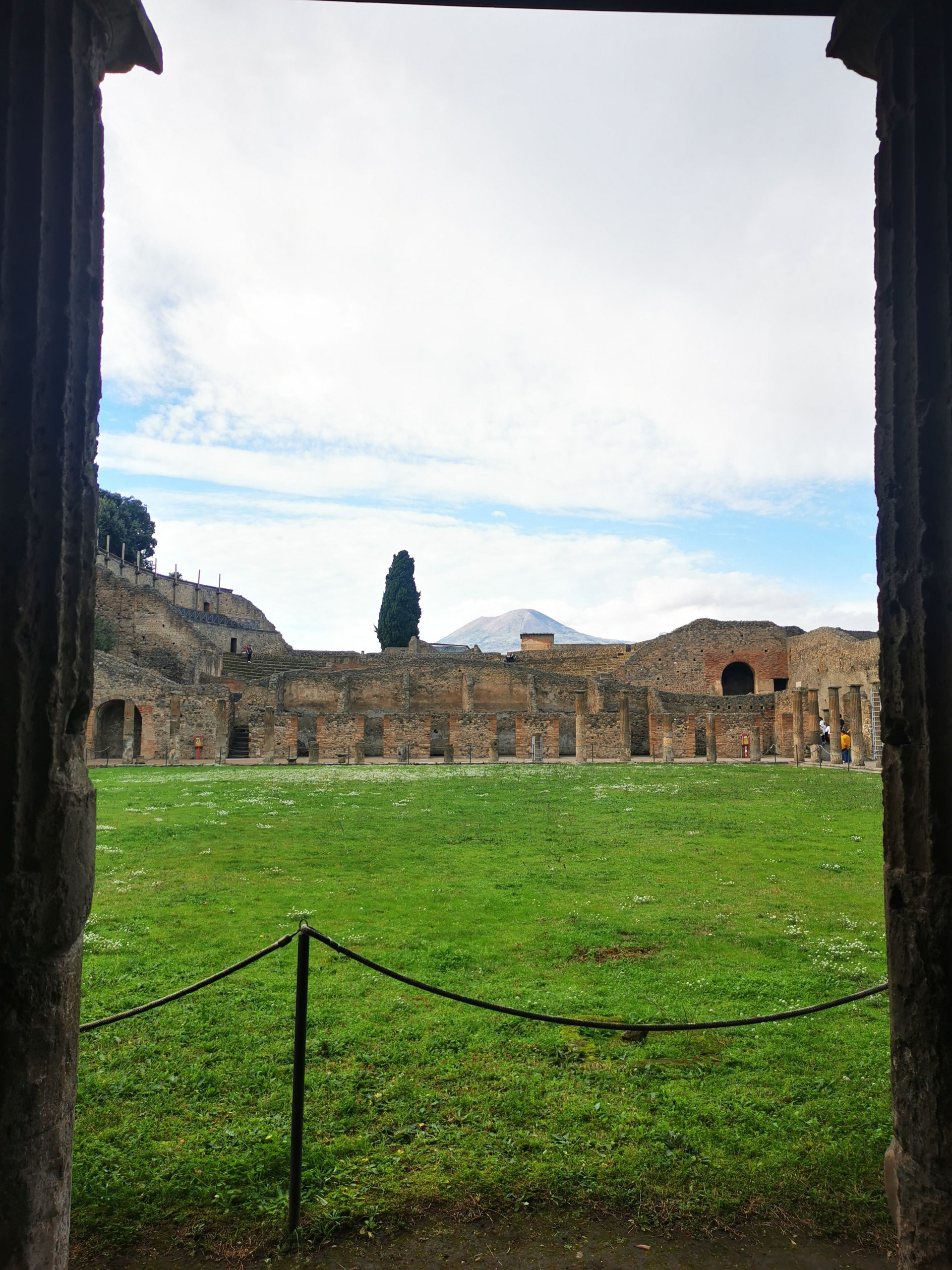 View through a dark doorway onto a grassy courtyard surrounded by arcades, with a distant volcano