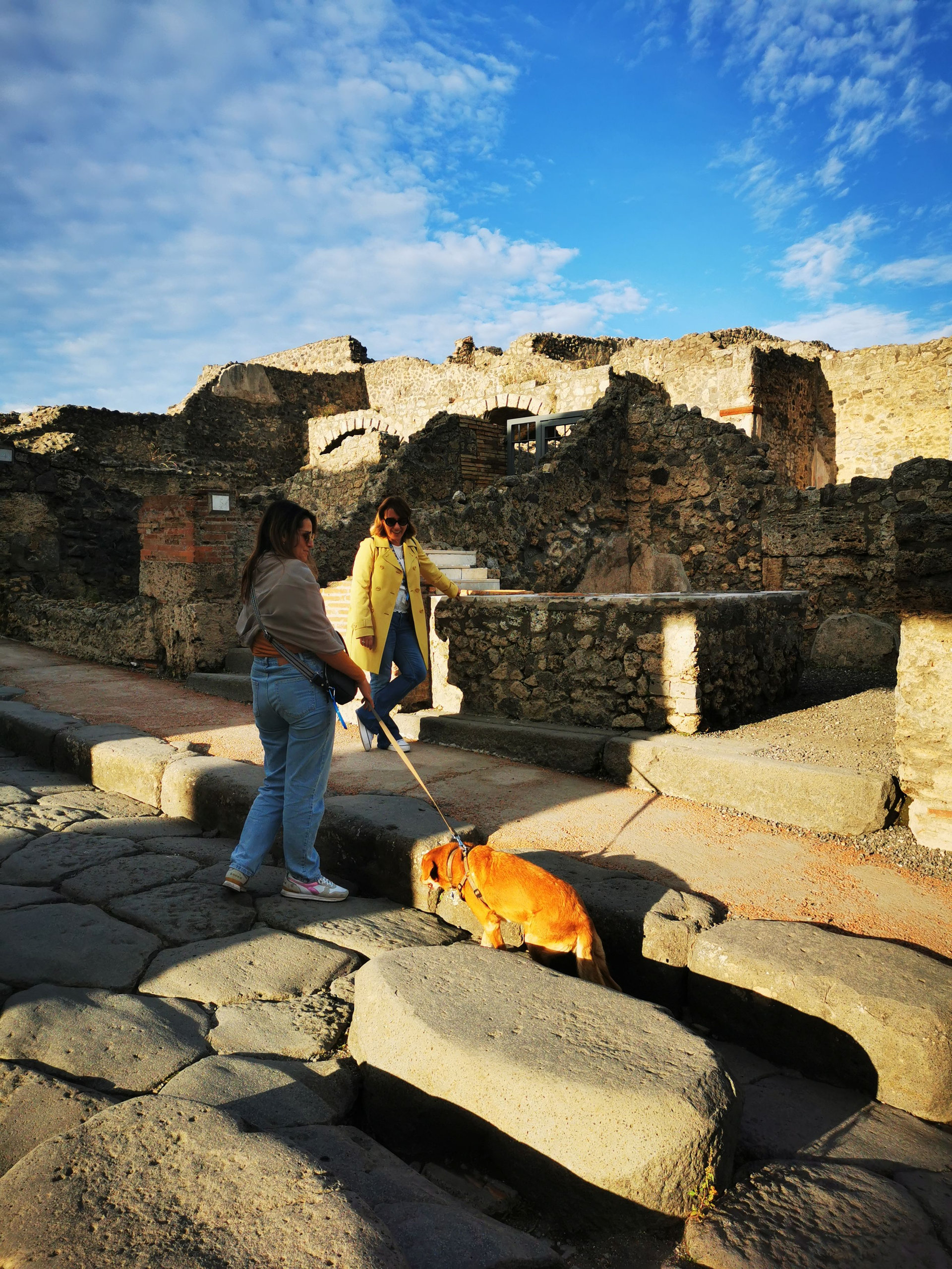 Two women walking a dog past large stepping stones in the ruins of an ancient town