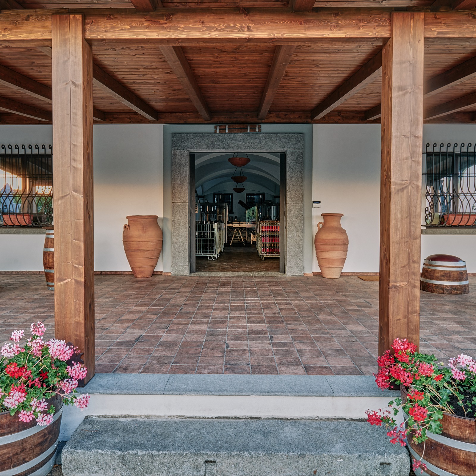Covered winery entrance with timber beams, terracotta amphorae and barrels of red geraniums