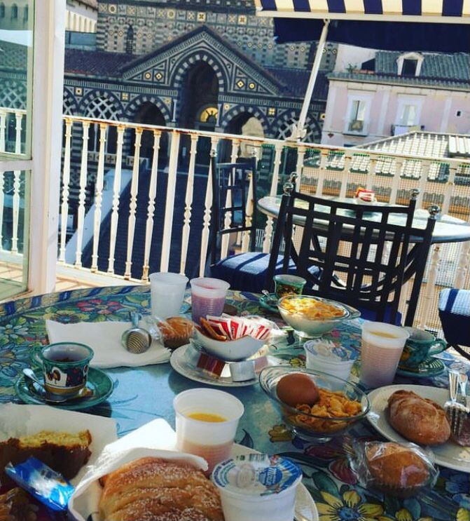 Breakfast of pastries, coffee and juice on a tiled table with a striped Amalfi cathedral facade behind