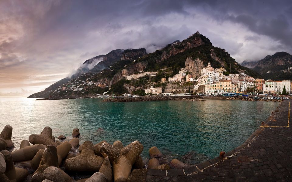 Amalfi seen across the water under a stormy sky, with concrete breakwater blocks in the foreground