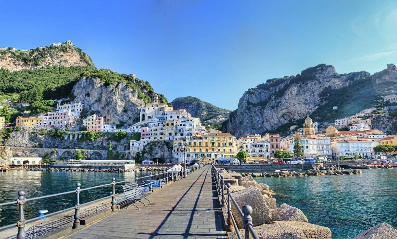 Pier leading toward Amalfi's cliffside houses and bell tower between mountains and sea