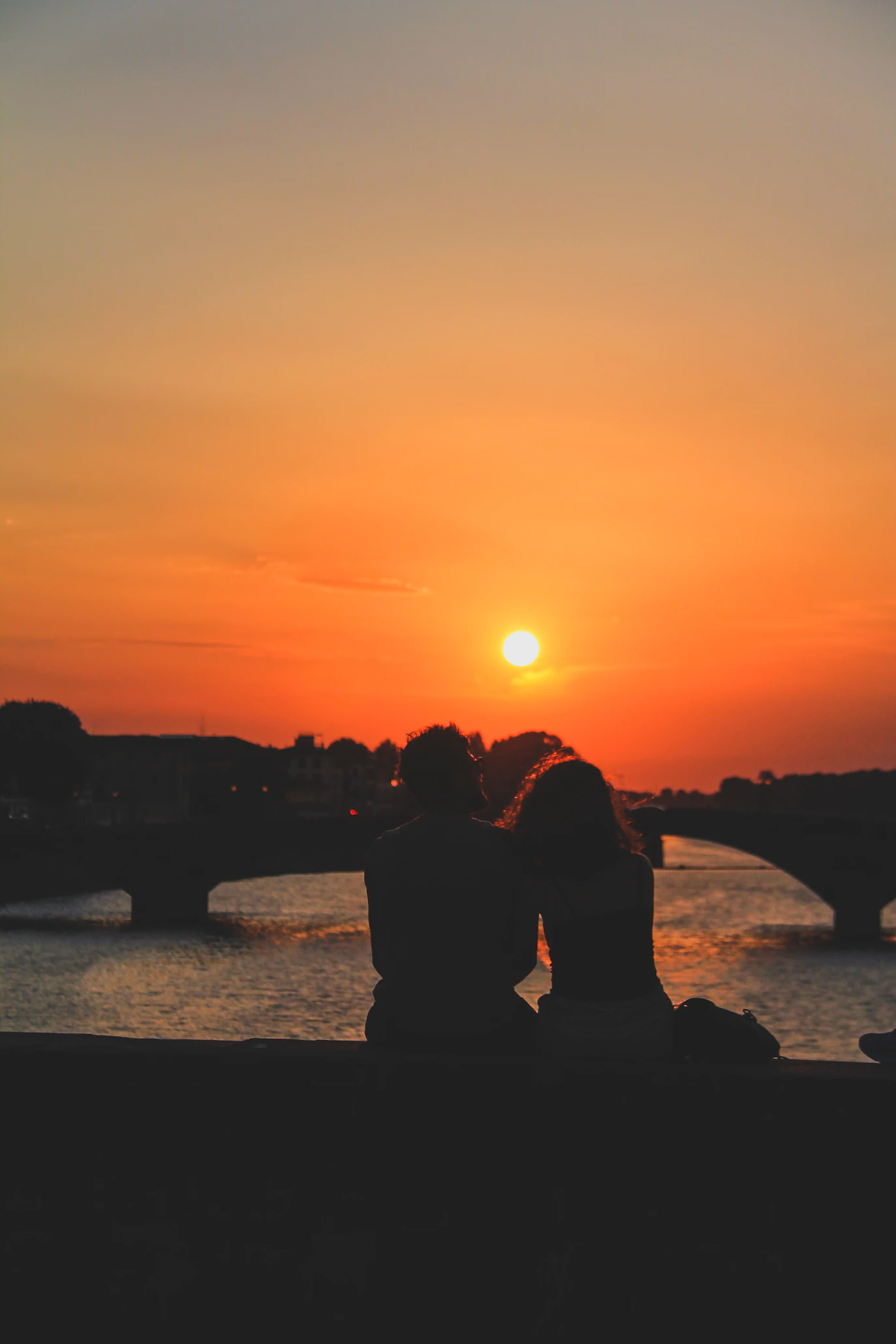 Couple silhouetted against a glowing orange sunset above an Arno bridge