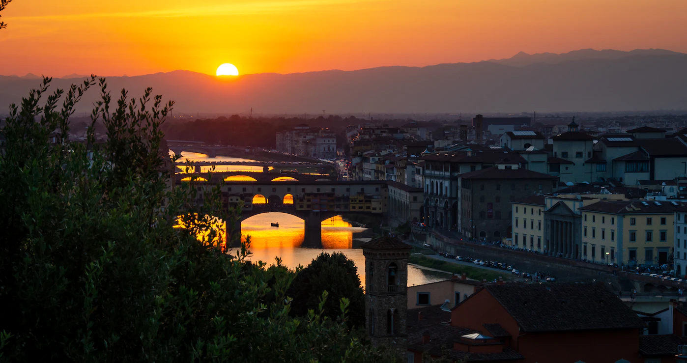 Sun setting behind hills over the Arno and Ponte Vecchio, seen through dark foliage
