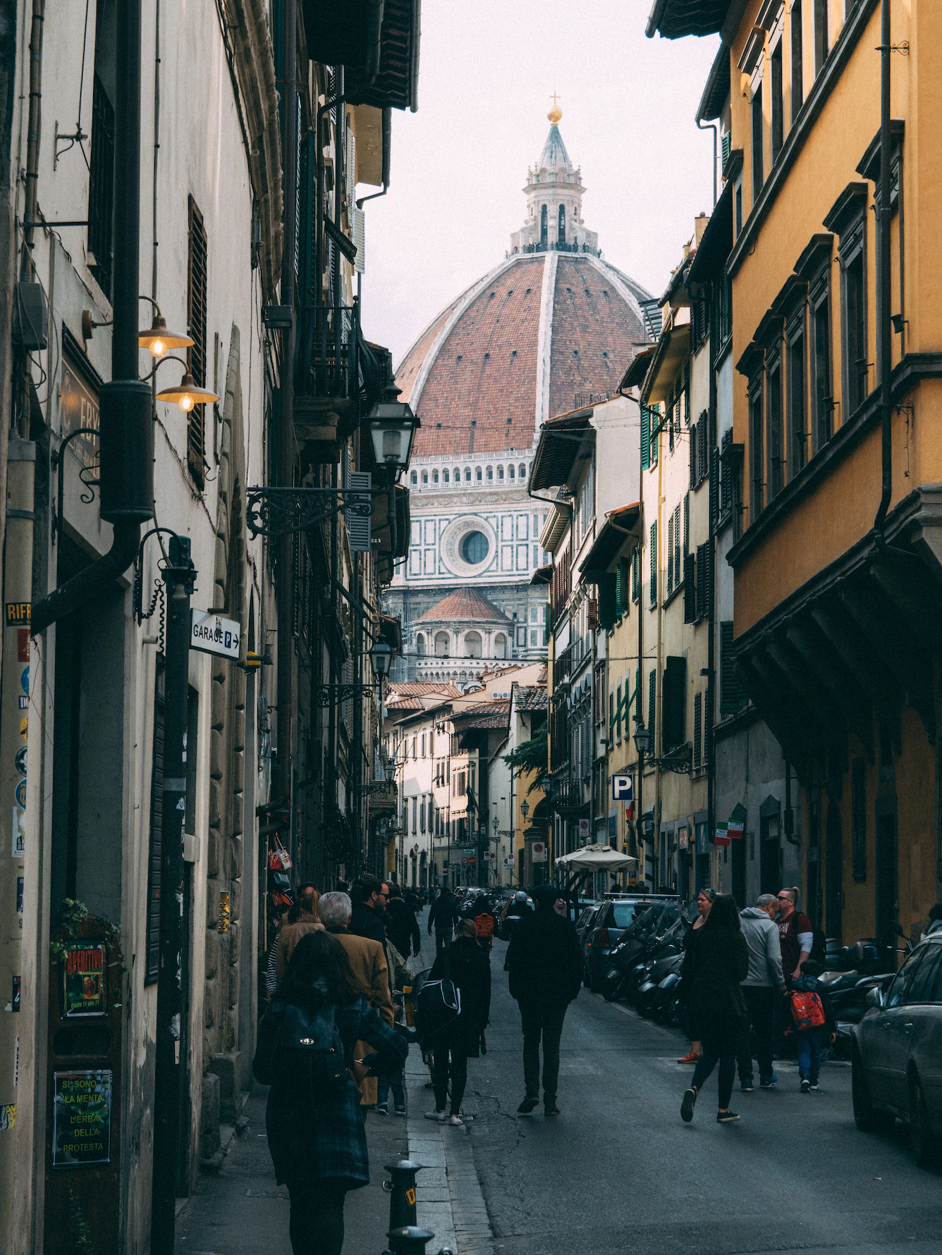 Narrow Florentine street with pedestrians and the terracotta dome of the Duomo at the end