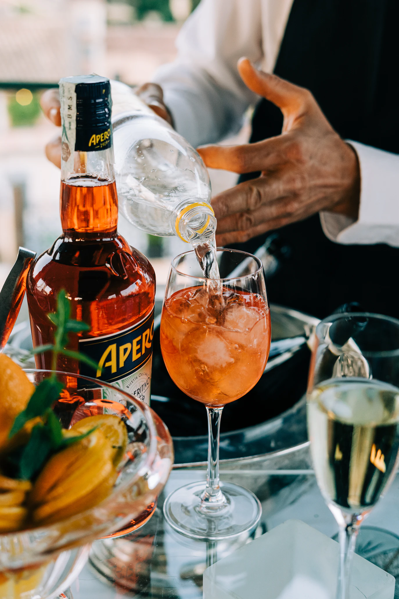 Aperol spritz being poured into a wine glass beside an Aperol bottle and a bowl of orange slices