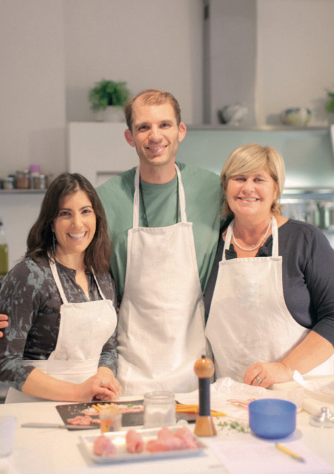 Three smiling cooks in white aprons at a kitchen counter with sliced ingredients before them