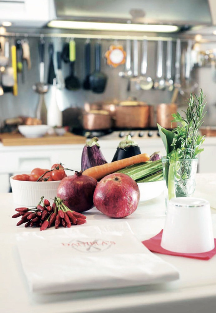 Fresh produce on a kitchen worktop, including pomegranates, aubergines, carrot, tomatoes and chillies, with copper pans behind