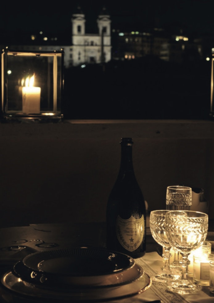 Candlelit dinner table set with crystal glasses and a champagne bottle, with a lit church façade in the night beyond
