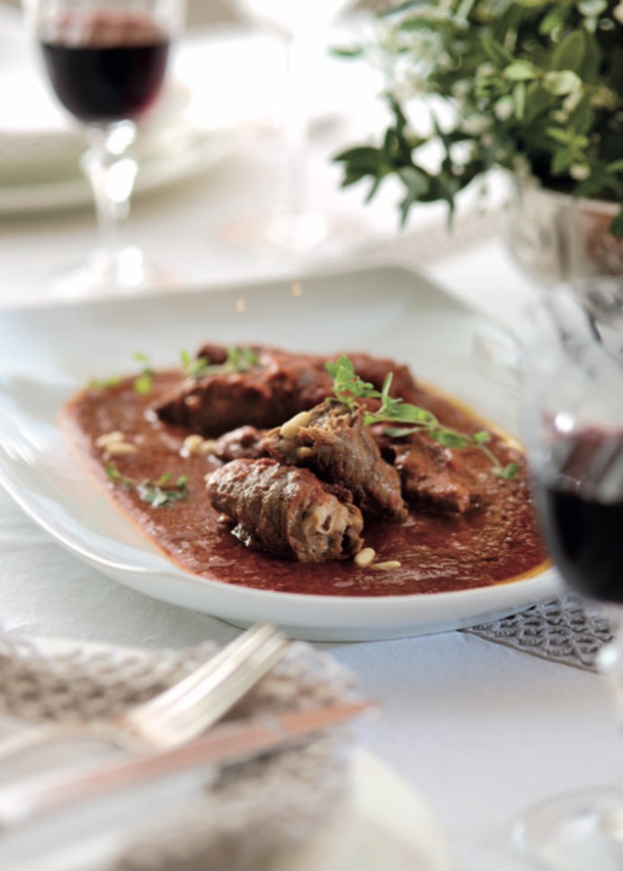 Beef rolls in tomato sauce garnished with herbs and pine nuts on an oval plate, with red wine glasses behind