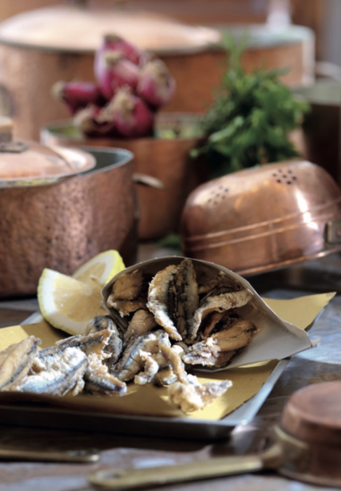 Paper cone of crispy fried anchovies with lemon wedges, set among copper pots