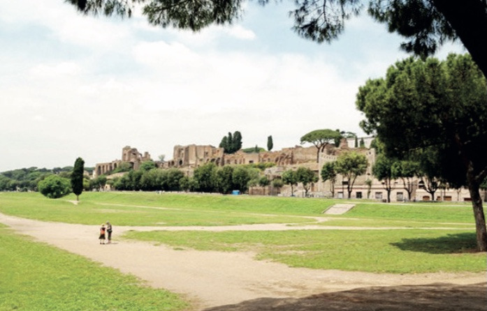 Wide grassy field of the Circus Maximus beneath the ruins of the Palatine Hill, shaded by umbrella pines