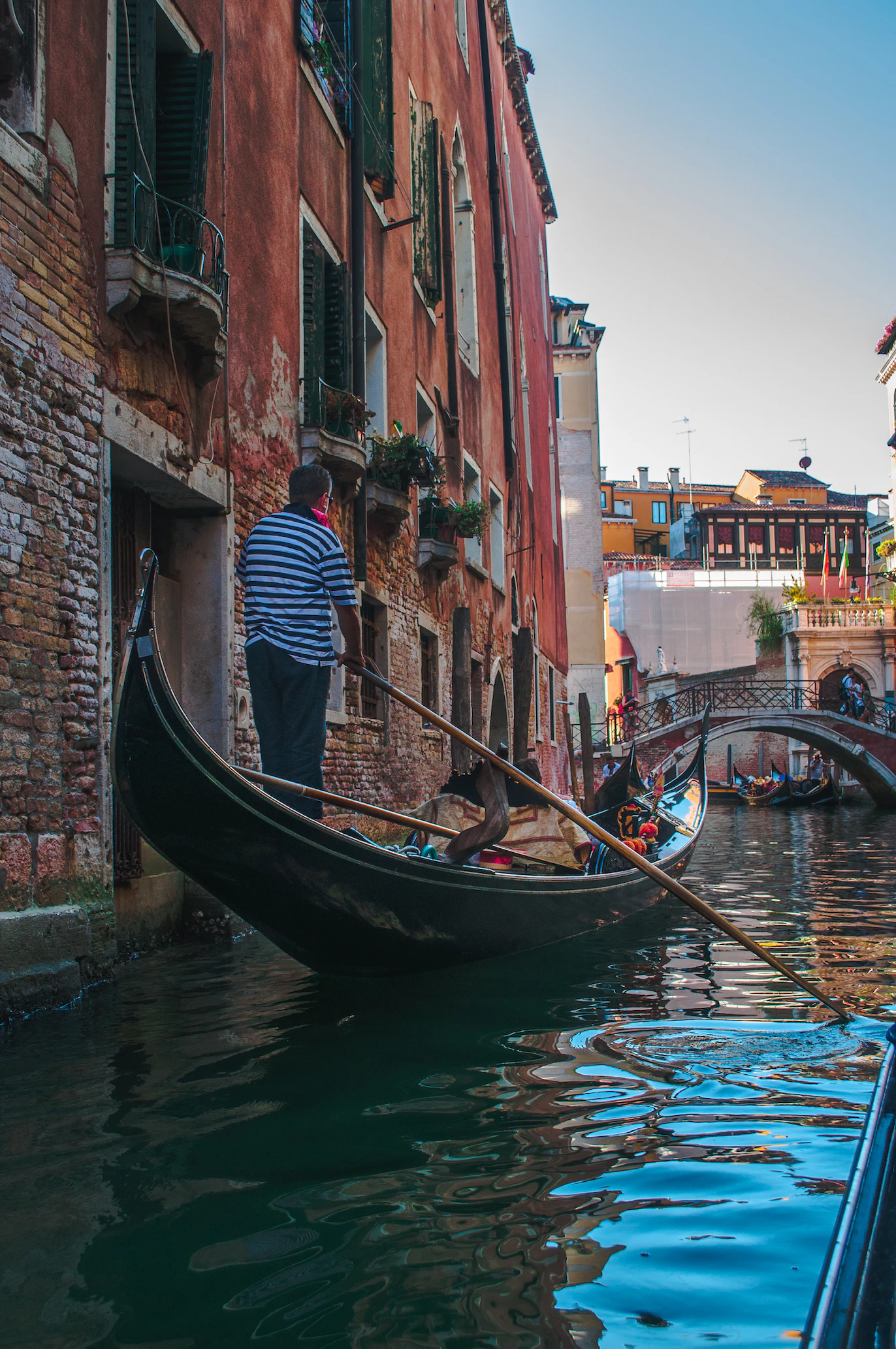 Gondolier in a striped shirt steering a gondola along a narrow canal beside brick palazzi
